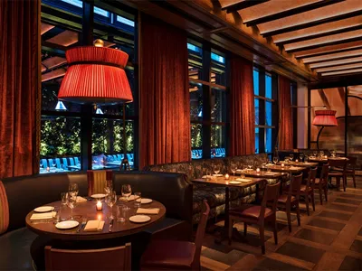 A luxurious dining room with red velvet curtains and wooden flooring, featuring round tables set for dinner with wine glasses