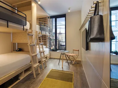 A minimalist hotel room with bunk beds, a foldable chair, a small table, and a potted plant.