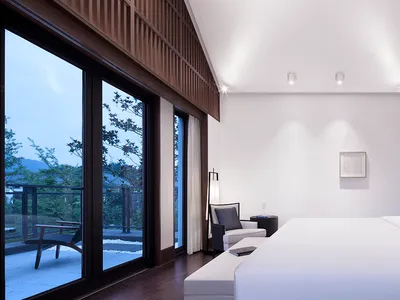 Minimalist-style bedroom featuring a large bed with a white sheet and dark wooden accents, with a view of the surrounding tre