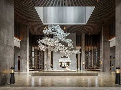 A modern hotel lobby featuring a large, metallic tree sculpture on a stone pedestal with tall columns and a skylight.