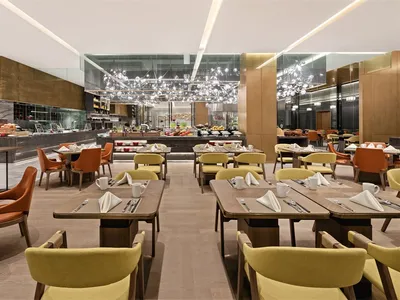 A modern restaurant interior featuring tables and chairs with wooden tops, orange and yellow upholstered chairs, and metallic