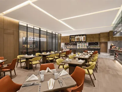 A spacious, modern restaurant interior with wooden tables, yellow chairs, and a well-lit kitchen area.