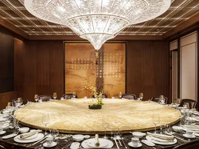 A grand dining room with a large, round marble table surrounded by elegant chairs. The centerpiece features a beautiful flowe