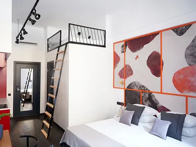 A modern minimalist bedroom with a bed, pillows, and abstract wall art. A loft area with a ladder staircase leads to another