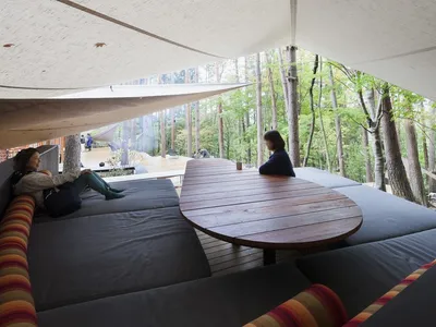 A modern, industrial-style lounge area nestled within a forest, featuring cozy sofas and a wooden table on a wooden floor, wi