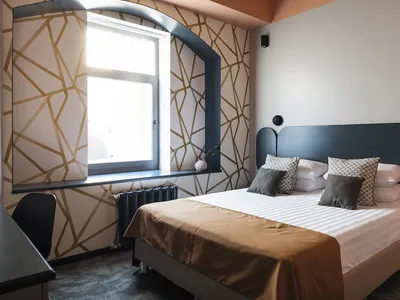 A modern, minimalist bedroom with geometric patterns on the wall and a bed with white and brown linens, under a large window.