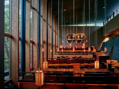 A modern restaurant interior featuring wooden tables and chairs, with hanging lights and a large window showing natural light