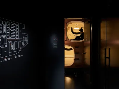 極簡臥室內，一人坐在膠囊旅館房間裡使用智慧型手機。