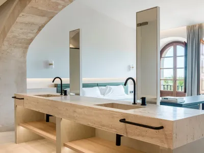 Modern minimalist hotel bathroom with marble countertops, sinks, and mirrors, featuring a king-sized bed with white bedding i