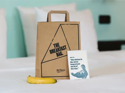 棕色紙袋標有「The Breakfast Bag」字樣，旁邊擺放香蕉，背景為臥室，另有一張印有引言的小卡片