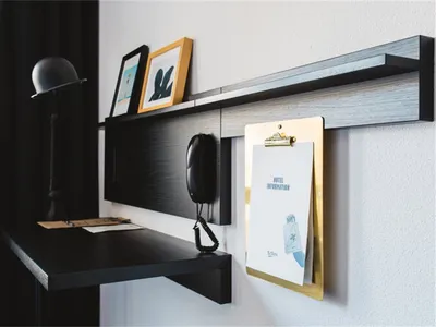 A minimalist hotel room desk with a black desk, black lamp, and a hanging hotel information clipboard.