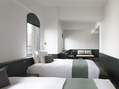 A minimalist hotel room with white walls and neutral-colored bedding, featuring a modern bench and minimalist decor.