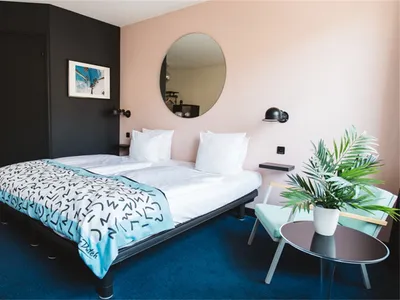 Modern hotel room with blue carpet, a double bed with white sheets and blue accent blanket, a small table, a green plant, and