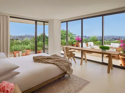 Modern bedroom with balcony overlooking cityscape, featuring a wooden bed frame, glass window, and fabric textiles.