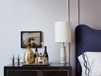 A modern bedroom corner with a black sideboard featuring a brass pineapple ice bucket, a framed artwork, a table lamp, and a