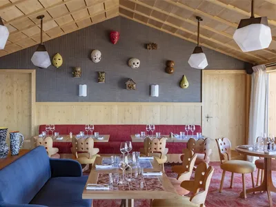 A modern restaurant interior featuring wooden furniture, red banquettes, and decorative wall masks.