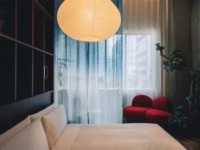 Modern minimalist hotel room with white textiles, red furniture, and a large window with blue curtains.