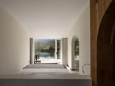 Minimalist bedroom with concrete walls and floor, framed by large windows overlooking a scenic lake view.