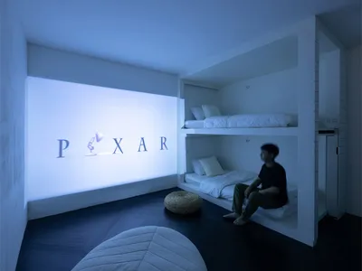 A minimalist living room featuring white walls, floor, and furniture, with a person sitting on a built-in bed under a Pixar l