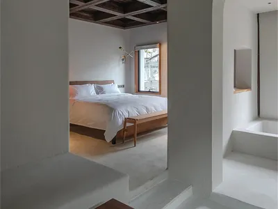 A minimalist bedroom with a concrete wall and floor, featuring a white bed and wood floor.