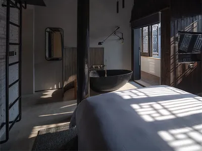 Interior of a minimalist bedroom with concrete walls and ceiling beams, featuring a black bathtub and a bed.