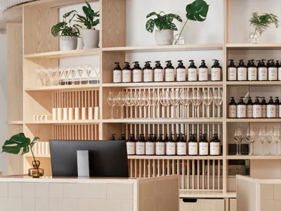 Modern restaurant interior with a wooden and tiled reception counter, glassware, and shelves with bottles and green plants.