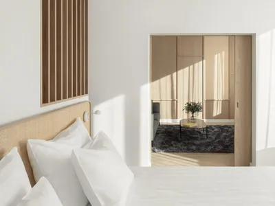 Modern, minimalist bedroom with white bedding, wooden furniture, and light streaming through the window.
