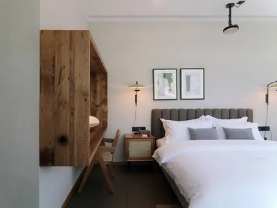 A minimalist bedroom featuring a wooden desk, concrete wall, and white textiles on a large bed.
