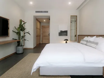 Modern minimalist bedroom featuring a white bed with wooden accents and fabric headboard against a plain wall, with a small p