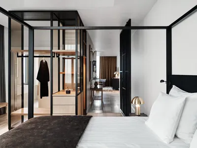 A minimalist bedroom with a bed, white bedding, and a glass-enclosed closet with wood shelves and metal accents.
