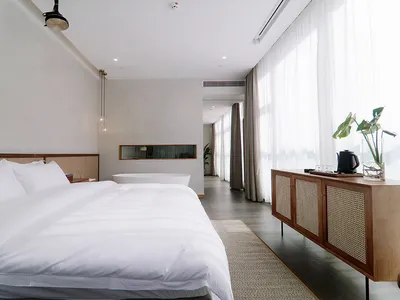 A modern minimalist bedroom with a white bed, wooden cabinet, and large windows.