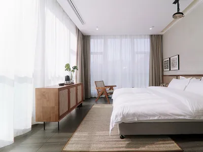 A modern, minimalist bedroom featuring a large window with white sheer curtains, a white bed with a beige headboard and white