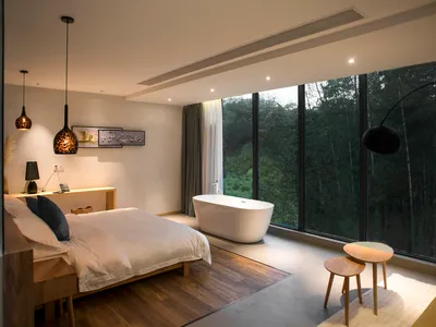 A modern minimalist bedroom featuring white bedding, wooden furniture, and a freestanding bathtub in front of a large window