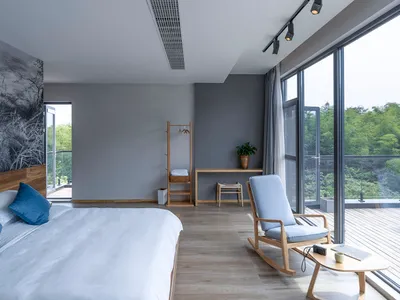 A minimalist bedroom with a large bed, wooden rocking chair, and floor-to-ceiling glass window overlooking greenery.