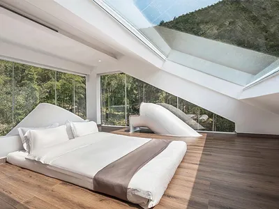 Modern minimalist bedroom with a bed, glass walls, and wooden flooring overlooking a forested area.