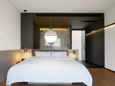 一間極簡臥室，白色床鋪靠著混凝土牆，搭配木質地板與吊燈燈具。