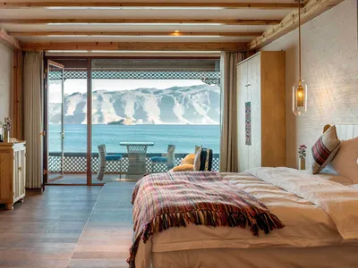 Modern hotel suite with wooden floor, ceiling, and furniture, featuring a balcony overlooking a lake and mountainous landscap