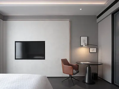 A minimalist bedroom featuring a black leather chair, a flat-screen television mounted on the wall, and a small round table w
