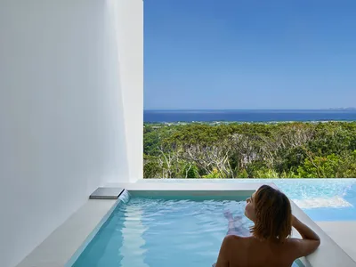 Person relaxing in a modern, minimalist hot tub overlooking lush greenery and the ocean.
