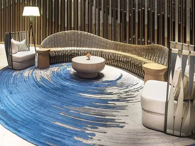 A modern hotel lobby featuring a curved, wicker sofa, a round wooden table with a floral centerpiece, and a circular blue rug