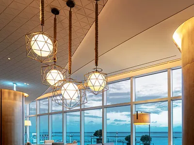A contemporary hotel lobby with wooden furniture, geometric lighting, and a view of the ocean.