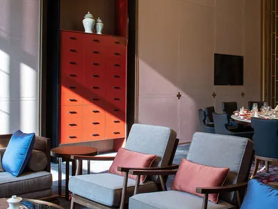 Modern interior design of a hotel lounge with grey armchairs, blue and pink cushions, and a wooden side table with a small te