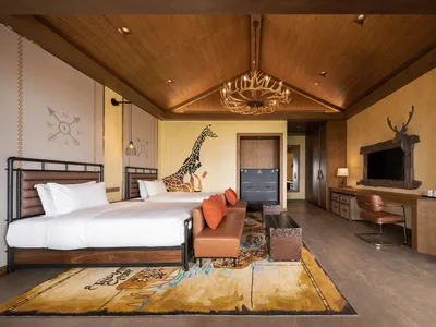 Modern hotel room featuring wood-themed decor and furniture, including a bed, sofa, desk, and chandelier.