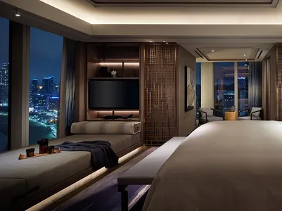 A contemporary bedroom featuring a large bed with white bedding, a seating area with a TV and lounge chairs, and a view of a