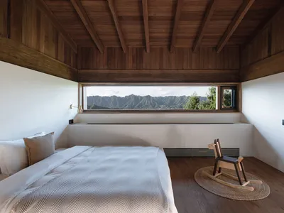 A minimalist bedroom with a large window overlooking mountains, featuring white walls, wood floors, a bed with a white comfor