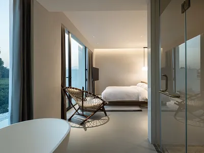 Modern minimalist bedroom with a wooden bed, glass walls, and fabric seating area overlooking greenery.