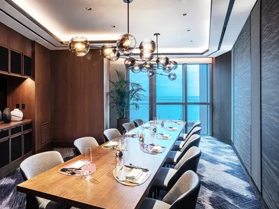 A modern restaurant dining room featuring a wooden table and chairs, chandelier, and ocean view.
