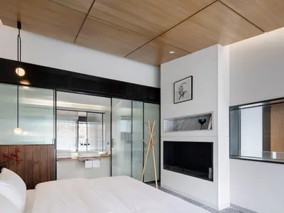 A minimalist bedroom featuring white bedding and lighting, with a glass-enclosed bathroom and wooden ceiling panels.