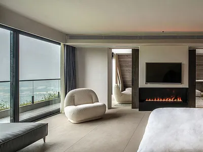 Modern minimalist bedroom featuring a large bed with white bedding, a glass fireplace, and large windows with glass walls.