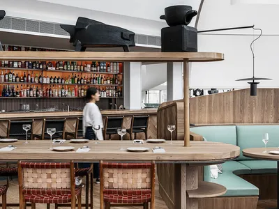 Industrial-style restaurant interior featuring wooden tables and chairs with leather seats, metal light fixtures, and a bar w